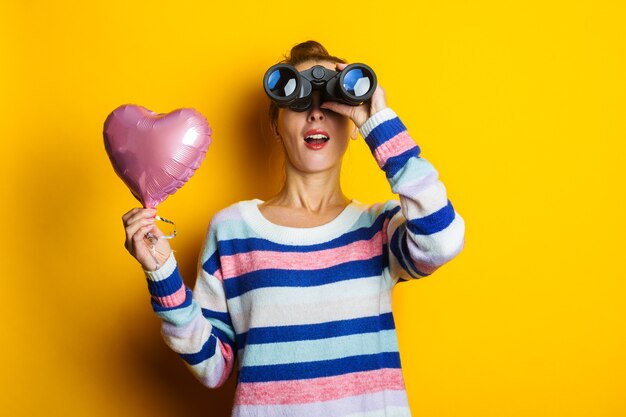 Young woman in a sweater holds an air balloon heart and looks through binoculars on a yellow background. Valentine's day composition.