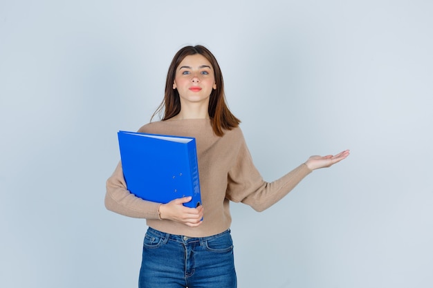 Young woman in sweater, jeans holding folder, spreading arm and looking pleased , front view.