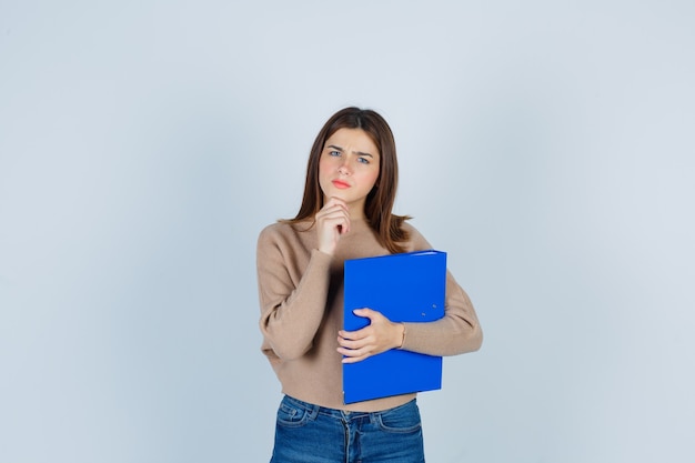 Young woman in sweater, jeans with hand on chin, keeping folder and looking puzzled , front view.