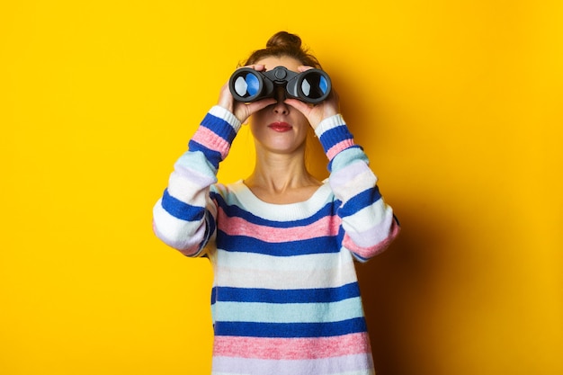 Young woman in sweater looking through binoculars on yellow background.