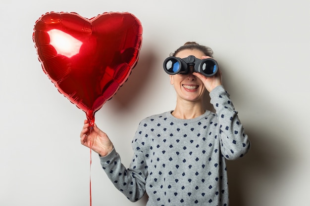 Giovane donna in un maglione guarda attraverso il binocolo e tiene un cuore di mongolfiera su uno sfondo chiaro. concetto di san valentino. cerca una persona cara.