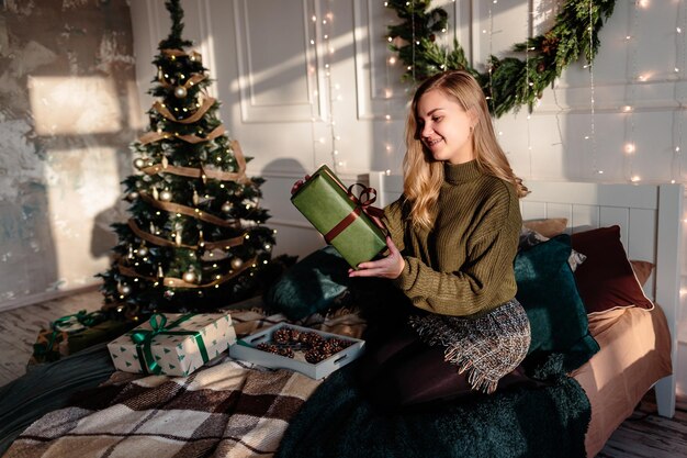 A young woman in a sweater opens Christmas gifts in a bedroom decorated for Christmas against the backdrop of a Christmas tree.