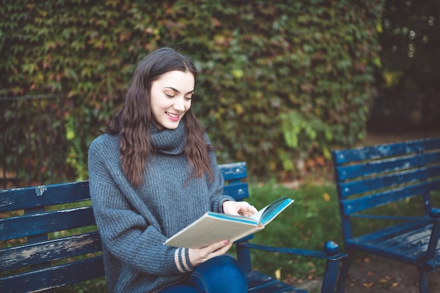 Young woman in a sweater reading a book, sitting on the bench in the park. A