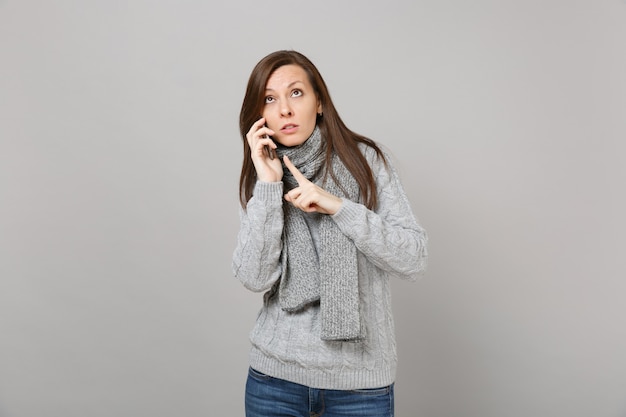 Young woman in sweater scarf pointing index finger, looking up, talking on mobile phone isolated on grey background. Healthy fashion lifestyle people emotions, cold season concept. Mock up copy space.