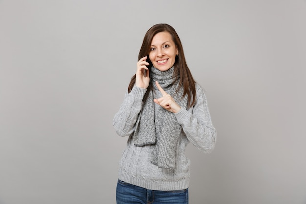 セーターを着た若い女性、灰色の背景で隔離の楽しい会話を行う携帯電話で話している人差し指を指すスカーフ。健康的なファッションライフスタイルの人々の感情の寒い季節のコンセプト。