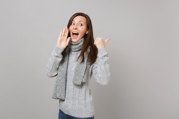 Young woman in sweater, scarf whispers gossip, tells secret with hand gesture, showing thumb aside isolated on grey background. Healthy fashion lifestyle, people sincere emotions, cold season concept.