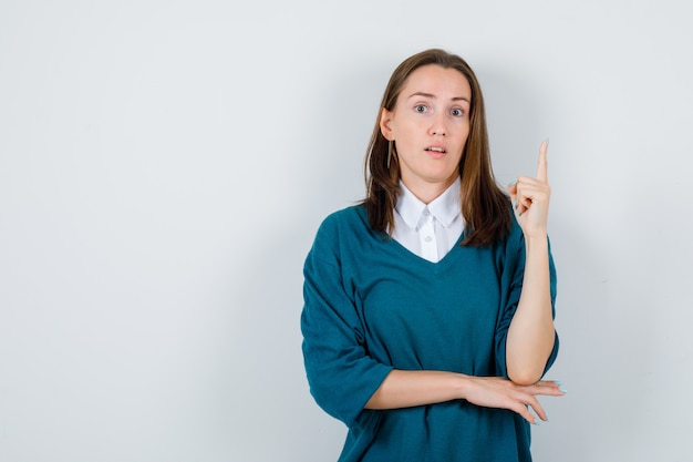 Young woman in sweater over white shirt pointing up and looking puzzled , front view.