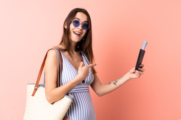 Young woman in swimsuit in summer holidays holding a passport over isolated pink background and pointing it