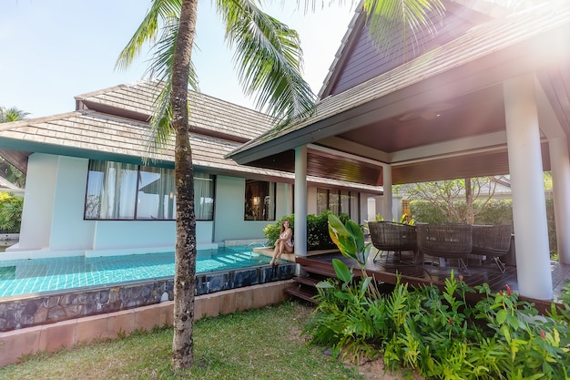 Young woman in swimwear sitting near swimming pool on tropical luxury villa