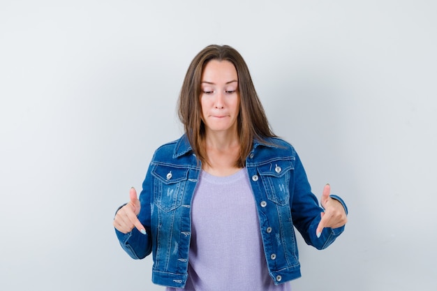 Young woman in t-shirt, jacket pointing down and looking curious , front view.