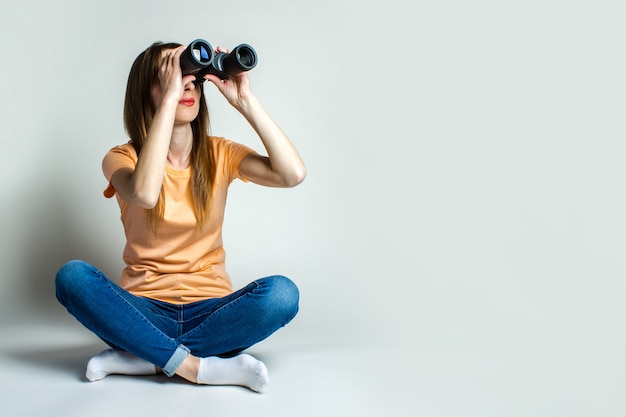 Young woman in a t-shirt and jeans sitting on the floor looks through binoculars on a light background