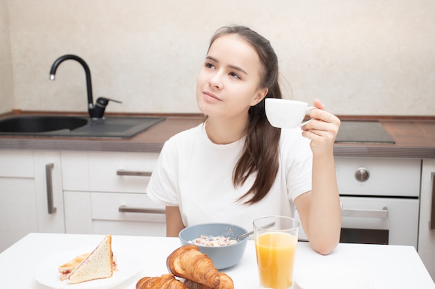 朝食を持っている台所のテーブルで若い女性はコーヒーを飲む