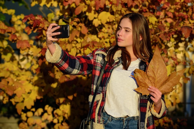 Giovane donna prendere selfie in autunno parco ragazza con telefono cellulare e foglie gialle foglia d'acero all'aperto f