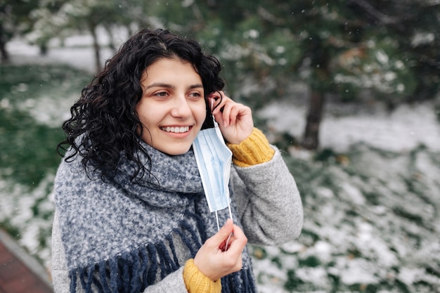 Young woman takes off the medical sterile mask at a winter snowy park on a cold frosty day.