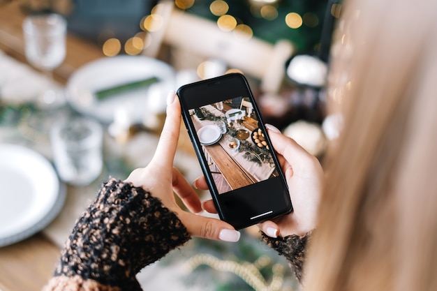 Young woman takes pictures of Christmas festive table on smartphone. Close-up