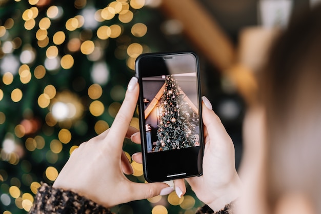 Young woman takes pictures of Christmas tree on smartphone. Close-up