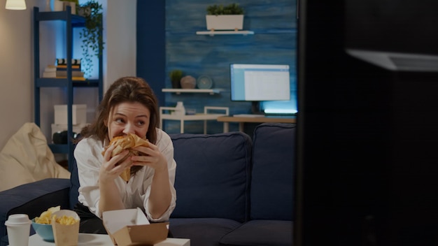 Young woman taking bite from hamburger sitting on couch while watching TV at home. Person eating takeaway meal from fast food restaurant while looking at television in living room