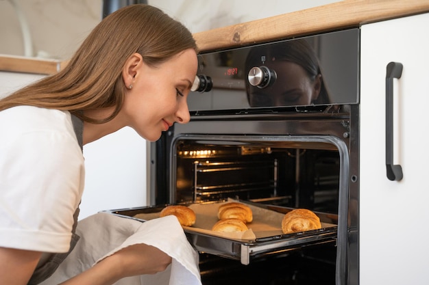 キッチンで上から焼きたてのクロワッサンのトレイを取り出す若い女性