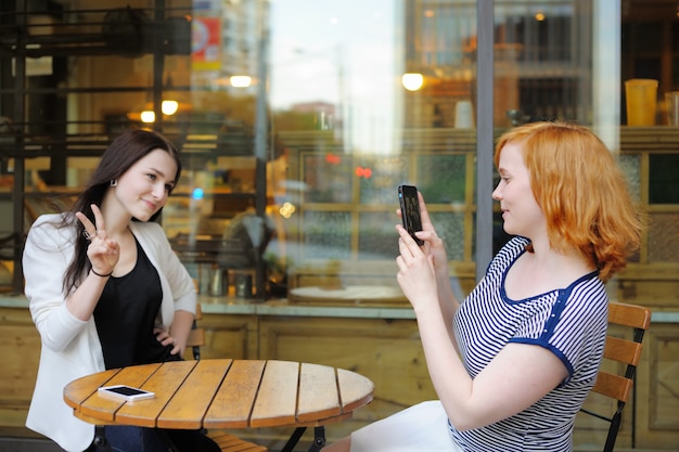 屋外カフェで彼女の友人（携帯に焦点を当てる）の写真を撮る若い女性