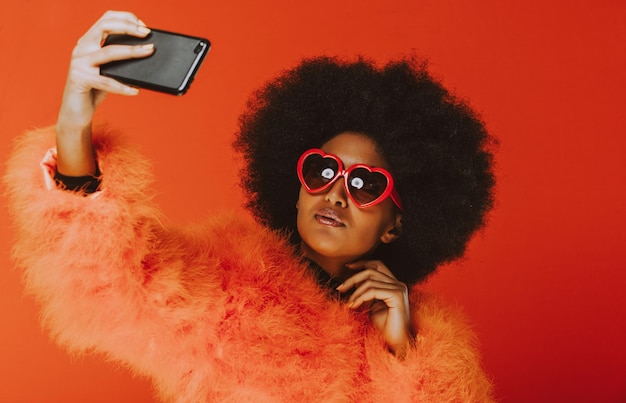 Young woman taking selfie on phone while wearing sunglasses against orange background