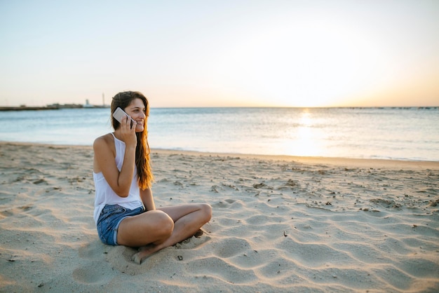 Молодая женщина разговаривает по мобильному телефону на пляже на закате