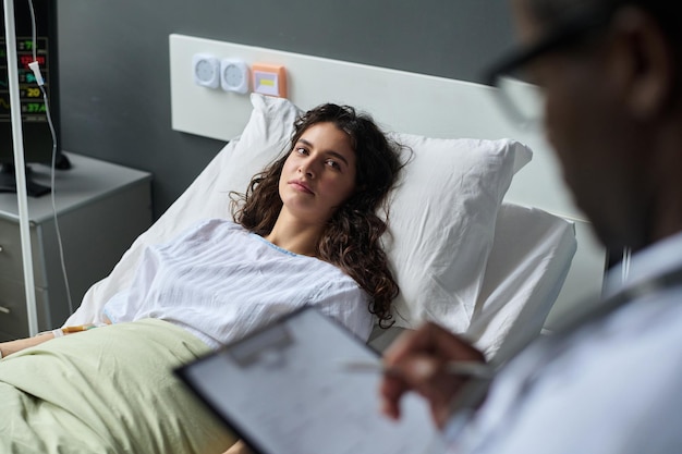 Young woman talking to doctor about her health condition while lying on bed in ward