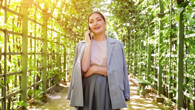 Young woman talking on the phone in an arched corridor of trees in the garden, sunlight