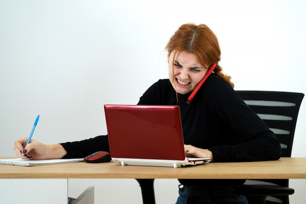 Young woman talking on a phone sitting at a desk with laptop and notebook.