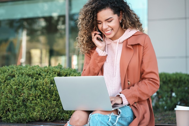 Young woman talking on the phone and using a laptop while sitting outdoors at the street. Urban concept.