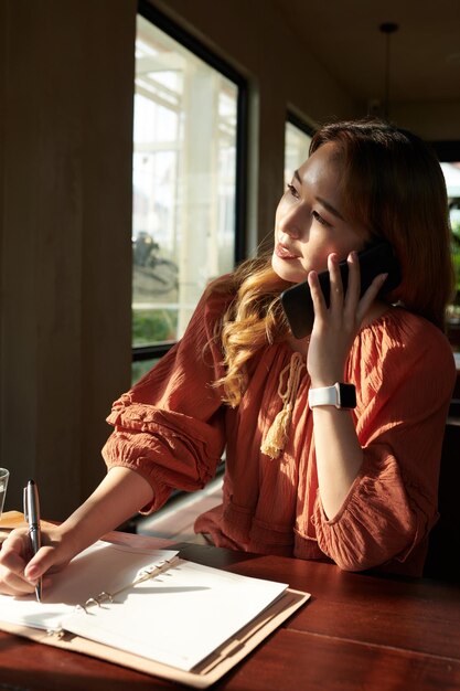 Young woman talking on phone wirh colleague or friend when taking notes in planner