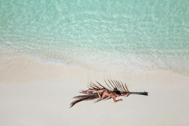 Photo young woman tanning sunbathing on palm tree leaf woman wearing bikini at the beach on a white sand from above view from drone