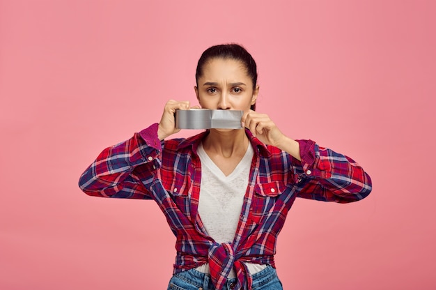 Foto la giovane donna si chiuse la bocca con il nastro adesivo, muro rosa, emozione. espressione del viso, persona di sesso femminile che guarda sulla fotocamera in studio, concetto emotivo, sentimenti