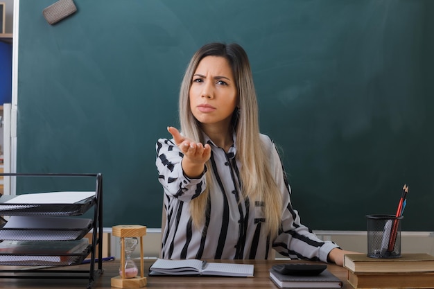 教室の黒板の前の学校の机に座っている若い女教師は、不機嫌に尋ねるように腕を上げている学生の宿題をチェックしています