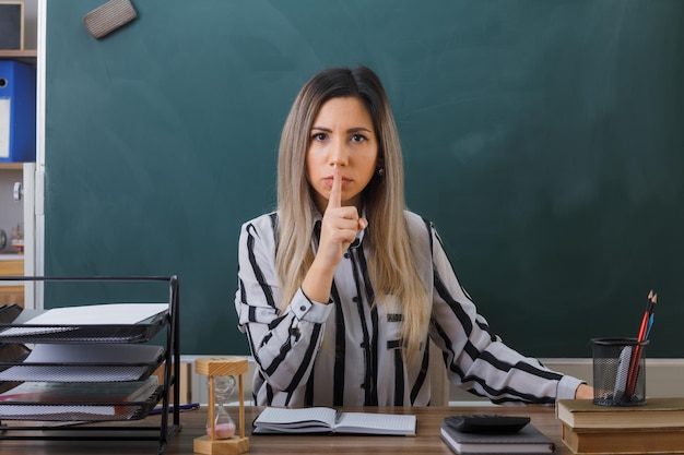 young woman teacher sitting at school desk in front of blackboard in classroom checking homework of students making silence gesture with finger on lips