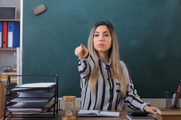 young woman teacher sitting at school desk in front of blackboard in classroom checking homework of students pointing with indexd finger at camera with serious face