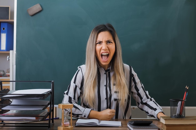 Young woman teacher sitting at school desk in front of blackboard in classroom checking homework of students shouting with aggressive expression angry and crazy mad clenching fist