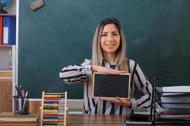 young woman teacher sitting at school desk in front of blackboard in classroom explaining lesson holding small chalkboard smiling confident happy and pleased