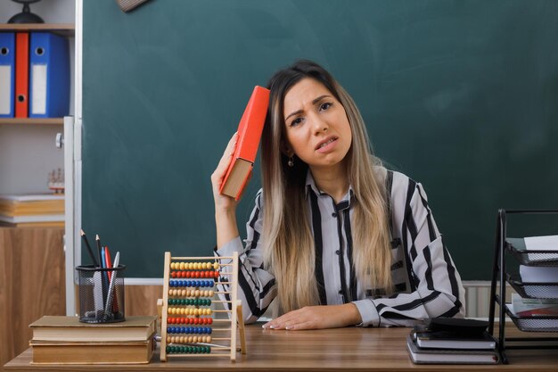 young woman teacher sitting at school desk in front of blackboard in classroom holding book looking tired and overworked