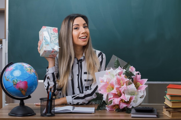 young woman teacher sitting at school desk in front of blackboard in classroom holding flower bouquet and present box from students looking happy and pleased happy teachers day concept