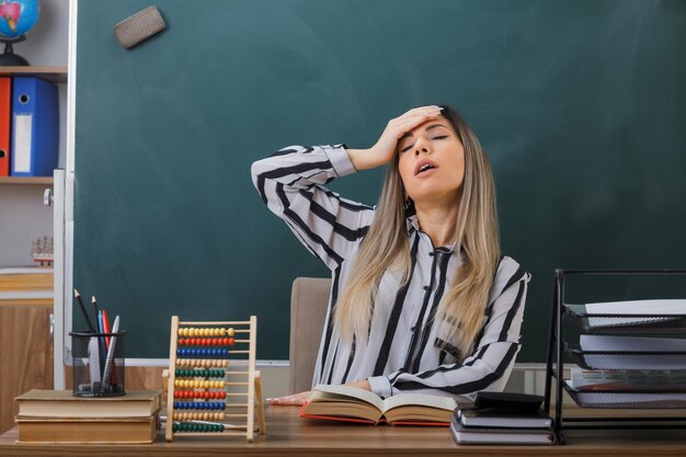 young woman teacher sitting at school desk in front of blackboard in classroom with book looking tired and overworked holding hand on her forehead