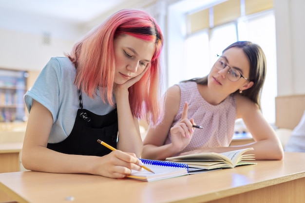 Photo young woman teacher teaches lesson in classroom of teenage children, teacher sits at desk with student, checks knowledge. education, school, college, teaching concept