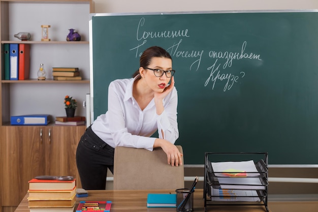 Young woman teacher wearing glasses explaining lesson looking confident and smart sitting at school desk in front of blackboard in classroom