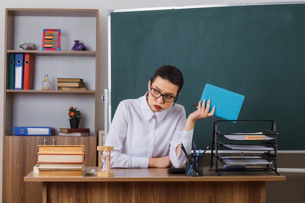 疲れて退屈そうな本を持って眼鏡をかけている若い女教師が教室の黒板の前の学校の机に座ってレッスンの準備をしている