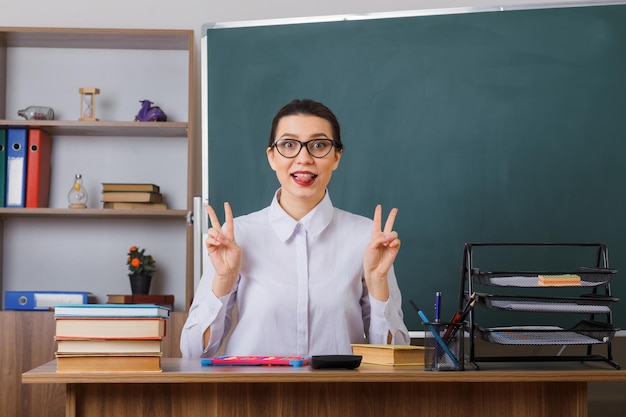 眼鏡をかけている若い女教師は、教室の黒板の前にある学校の机に座って舌を突き出しているvsignsを見せてうれしそうに見えます