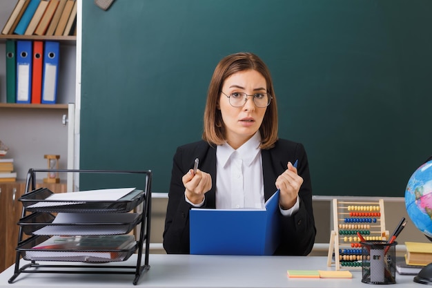 Young woman teacher wearing glasses sitting at school desk in front of blackboard in classroom checking class register pointing with pointer at camera being displeased