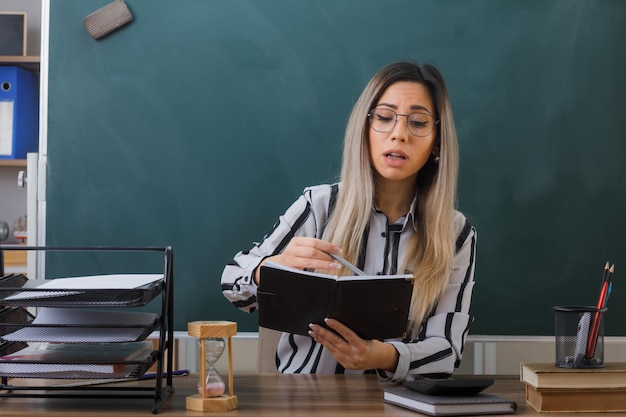 young woman teacher wearing glasses sitting at school desk in front of blackboard in classroom checking homework of students looking concentrated