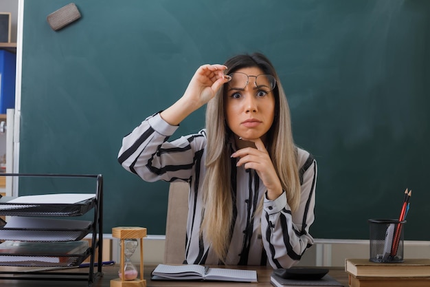 young woman teacher wearing glasses sitting at school desk in front of blackboard in classroom checking homework of students taking off glasses looking surprised and worried