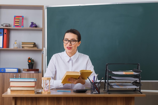 Young woman teacher wearing glasses sitting at school desk in front of blackboard in classroom explaining lesson smiling friendly