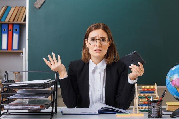 Young woman teacher wearing glasses sitting at school desk in front of blackboard in classroom using calculator preparing for lesson looking confused having no answer