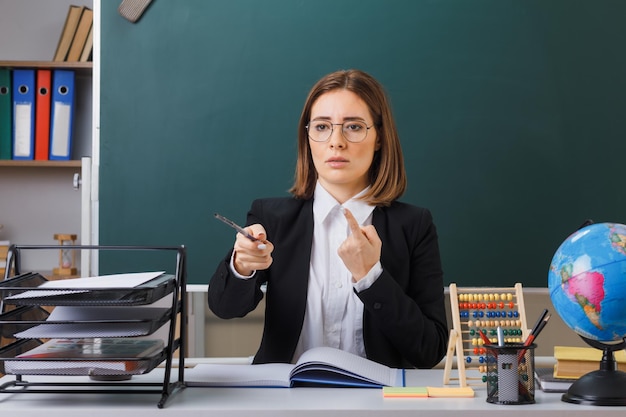 young woman teacher wearing glasses sitting at school desk in front of blackboard in classroom with abacus and globe checking class register holding pointer looking confident showing index finger